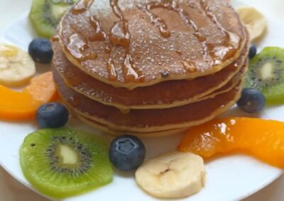 Stos naleśników z syropem, kiwi, bananem, mandarynką i jagodami na białym talerzu.