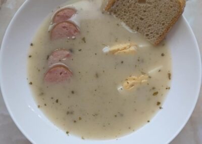 Talerz z żurkiem, kiełbasą i jajkiem, z kromką chleba na brzegu talerza.